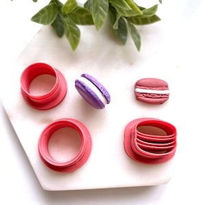 May include: A collection of pink macaron cookie cutters and two macarons. One macaron is purple with white filling, and the other is pink with white filling. The cutters are various shapes and sizes.