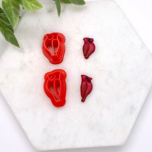 May include: Four red plastic cookie cutters, two are cardinal birds and two are cardinal shapes.