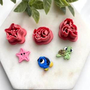 May include: A collection of pink and colorful polymer clay cutters and figurines. The cutters are in the shapes of a starfish, a rose, and a sea creature. The figurines include a pink starfish, a blue fish, and a green turtle.