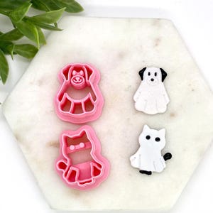 May include: Pink plastic dog and cat-shaped cookie cutters next to white dog and cat-shaped decorations. The cookie cutters are in the shape of a dog and a cat. The decorations are white with black accents.