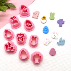 May include: A set of 14 pink plastic Easter cookie cutters, including a bunny, chick, cross, egg, and paw print. There are also several colourful fondant decorations, including a blue egg, a purple cross, a green bunny, a pink bunny, a white bunny, a white paw print, a yellow chick, and a blue bird.