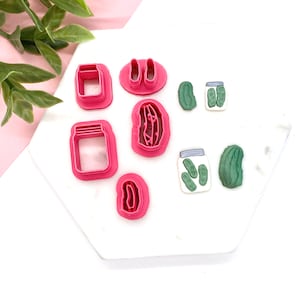 May include: A set of pink plastic cookie cutters shaped like jars, pickles, and a pickle jar. The cutters are on a white surface.