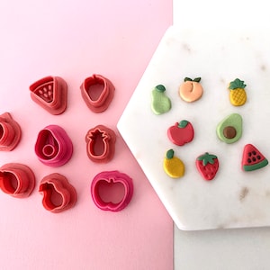 May include: A set of 9 pink fruit-shaped cookie cutters and 9 colorful clay fruit shapes on a white marble surface.