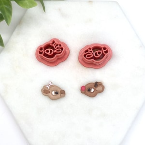 May include: Four pieces: two pink cookie cutters with the word "Joy" and two reindeer-shaped decorations. The reindeer decorations are brown with white accents, one with a pink nose and the other with a white bow.