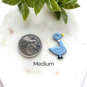 May include: A light blue pigeon-shaped charm with a yellow beak, white neck band, and black feet, positioned next to a Canadian 25-cent coin for scale. The word Medium is printed below the charm. The object is on a white marble surface.