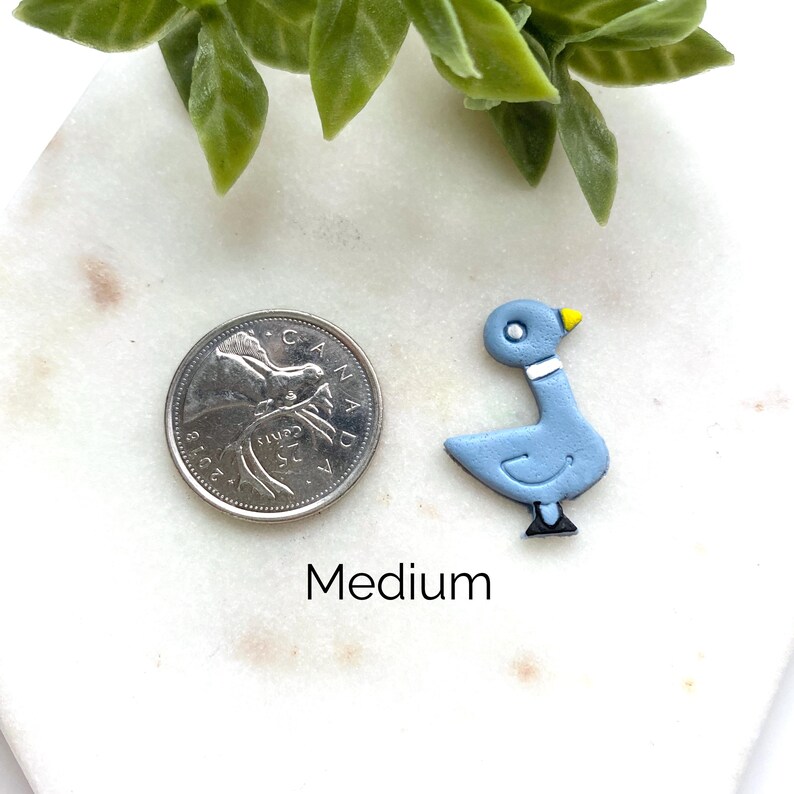 May include: A light blue pigeon-shaped charm with a yellow beak, white neck band, and black feet, positioned next to a Canadian 25-cent coin for scale. The word Medium is printed below the charm. The object is on a white marble surface.