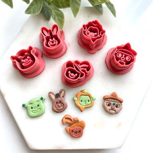 May include: A collection of pink plastic cookie cutters in various character shapes, including a pig, donkey, and a character with a hat. Below are several decorated cookies in the same character shapes, with green, brown, and orange details.