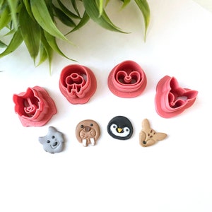 May include: A collection of pink plastic cookie cutters in various shapes, including a flower, a heart, and animal faces like a walrus, penguin, and fox. The cutters are arranged on a white surface with a green plant in the background.