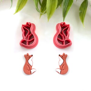 May include: Two pink fox-shaped cookie cutters and two fox-shaped embellishments. The cookie cutters have a detailed outline of a fox. The embellishments are orange with white and black accents. The items are on a white surface with green leaves.