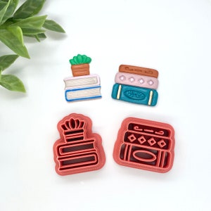 May include: Four cookie cutters shaped like stacks of books. Two are red and two are decorated with blue, green, and brown details. One of the red cutters has a small plant on top.