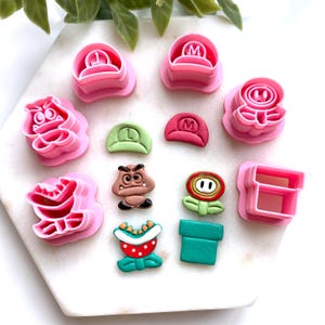 Peut inclure: Un ensemble d'emporte-pièces en plastique rose et de décorations en fondant colorées représentant des personnages et des éléments de la série de jeux vidéo Super Mario. Comprend des chapeaux de Mario, des Goombas et des fleurs.
