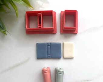 May include: A set of red plastic cookie cutters shaped like books. There are two different sizes of book cutters, a small square cutter, and two small book shapes made of fondant in pink and blue.