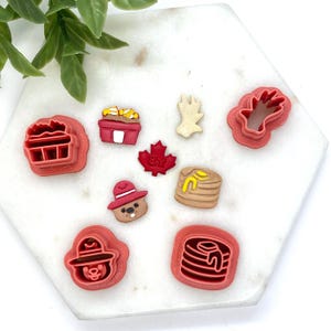 May include: A set of nine red plastic cookie cutters featuring Canadian themes, including a maple leaf, a beaver wearing a firefighter hat, a stack of pancakes with syrup, a poutine container, and a hand reaching for a maple leaf.