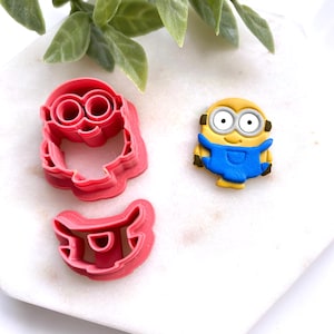 May include: A Minion-themed cookie cutter set and a Minion figurine. The cookie cutters are pink plastic and shaped like the Minion's face and overalls. The figurine is yellow with a blue jumpsuit and large eyes. The items are on a white surface.