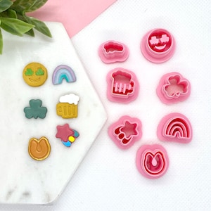 May include: A set of 10 pink plastic cookie cutters in various shapes, including a rainbow, a cloud, a beer mug, a shamrock, a horseshoe, a star, a smiley face, and a heart. The cutters are arranged on a white surface.