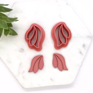 May include: Four pink plastic cookie cutters in the shape of flower petals. The cutters are on a white marble surface.