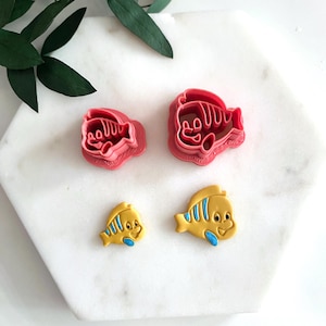 Puede incluir: Cuatro cortadores de galletas con forma de pez. Dos son rojos y dos son amarillos con detalles azules. Los cortadores rojos tienen la forma de un pez de dibujos animados con una boca grande. Los cortadores amarillos tienen la forma de un pez de dibujos animados con una boca más pequeña y una aleta en la espalda.