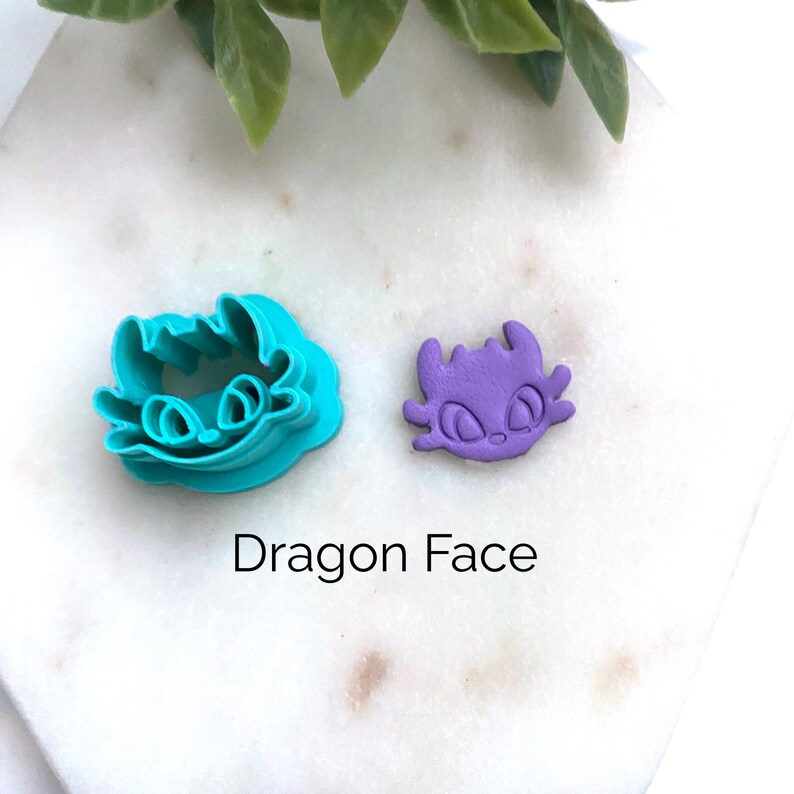 K&ouml;nnte beinhalten: Ein t&uuml;rkisfarbener Ausstecher aus Kunststoff in Form eines Drachengesichts und ein passendes, lila geformtes Drachengesicht aus Fondant. Das Design zeigt gro&szlig;e mandelf&ouml;rmige Augen, spitze Ohren und kleine H&ouml;rner. Der Text Dragon Face steht darunter.