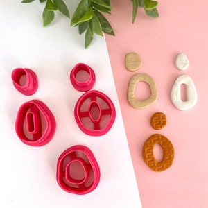 May include: A set of six pink polymer clay cutters in various shapes, including circles, ovals, and a ring shape. The cutters are arranged on a white surface. Next to the cutters are six polymer clay shapes in various colors, including brown, beige, and white.