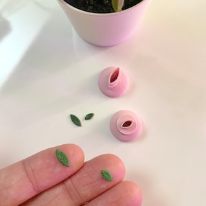 May include: Four small, green paper leaves and two pink paper flower buds. The leaves are in a hand, and the buds are on a white surface.