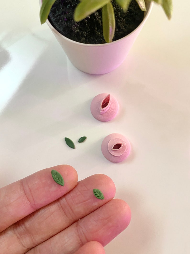 May include: Four small, green paper leaves and two pink paper flower buds. The leaves are in a hand, and the buds are on a white surface.