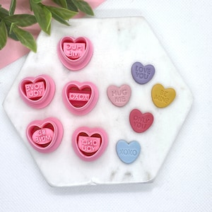 May include: A set of ten pink heart-shaped cookie cutters with various Valentine's Day messages, including "Hug Me", "Love You", "Be Mine", "Only You", "XOXO", and "You're Mine".