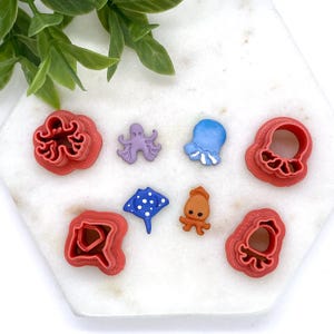 May include: A set of eight red plastic cookie cutters shaped like various sea creatures, including an octopus, a jellyfish, a stingray, and a starfish. The cutters are arranged on a white marble surface.
