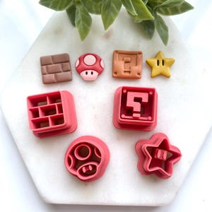 May include: A selection of colourful cookie cutters shaped like characters and objects from the Super Mario video game series. Includes a brown brick, a red and white mushroom, a question mark block, a yellow star, and other shapes.