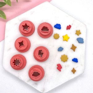 May include: A set of six red plastic cookie cutters with different shapes: a star, a cloud, a lightning bolt, a sun, a heart, and a cloud. There are also several small, colorful, decorative shapes on a white marble surface.