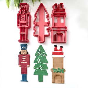 May include: A selection of Christmas cookie cutters. Features red plastic cutters of a nutcracker, Christmas tree, and house. Also, a decorated nutcracker, green tree with lights, and Santa in a chimney cookie.
