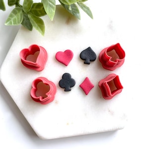 May include: A collection of playing card suit shapes, including hearts, spades, clubs, and diamonds. The set includes both the cut-out shapes and the corresponding cutters in red and black. The shapes are arranged on a white surface with a green plant in the background.