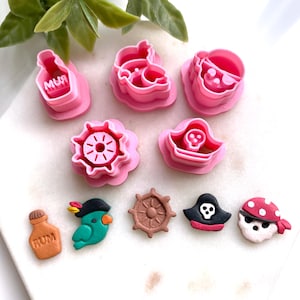 May include: A collection of pink plastic cookie cutters and decorative pirate-themed embellishments. The cutters include a bottle with the word "RUM", a parrot, a ship's wheel, and a skull and crossbones. The embellishments include a bottle of rum, a parrot, and a skull.