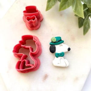 May include: Three red plastic cookie cutters in the shape of a hat and Snoopy. A Snoopy figurine is wearing a green hat and bow tie. The figurine is white with black ears and a black nose.