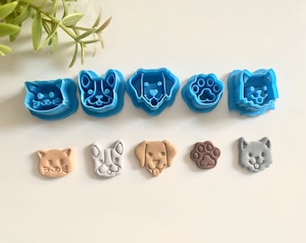 May include: A selection of blue plastic cookie cutters in the shapes of cats, dogs, and a paw print. Below are the corresponding biscuits in tan, white, brown, and grey.