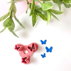 May include: A pink plastic butterfly-shaped cookie cutter with three distinct butterfly-shaped cutouts arranged in a triangular formation, accompanied by three small, solid blue butterfly-shaped pieces placed to the right of the cutter.