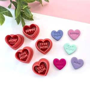 May include: A set of nine heart-shaped cookie cutters, with various messages including "Mom", "Dog Mom", "Cat Mom", "Best Mom", and "T238 Mom". The cutters are red and the hearts are in various colours, including purple, pink, blue, and green.
