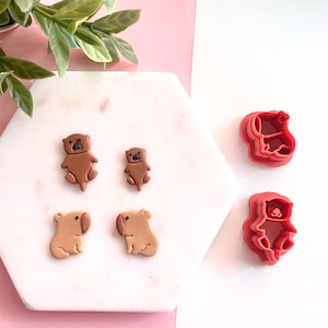May include: Two brown clay animal figurines of otters and two brown clay animal figurines of wombats, along with two red plastic cookie cutters shaped like otters and wombats.