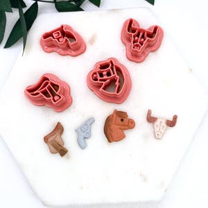 May include: A collection of cowboy-themed cookie cutters and decorative elements. The set includes pink plastic cutters in the shapes of a horse head, a bull skull, and a saddle. Also included are a light blue pistol and a brown horse head.