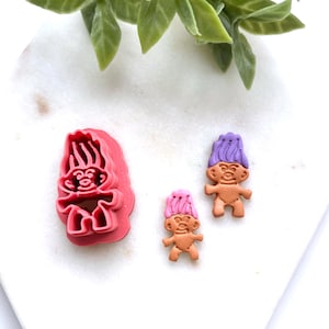 May include: A pink plastic cookie cutter shaped like a troll doll, alongside two troll doll-shaped sweets. One sweet has purple hair, the other pink. The sweets are tan with smiling faces.
