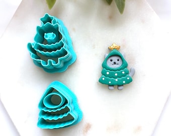 Könnte beinhalten: Zwei türkisfarbene Kunststoff-Ausstechformen in Form von gestuften Weihnachtsbäumen und eine kleine graue Katzenfigur, die ein grünes Weihnachtsbaumkostüm mit weißen Punkten und einem goldenen Stern an der Spitze trägt, auf weißem Untergrund.