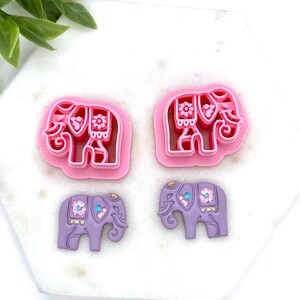May include: Two pink elephant-shaped cookie cutters and two lavender elephant-shaped embellishments. The cookie cutters have a detailed floral design. The embellishments have gold and turquoise accents, and floral details. The items are on a white surface.