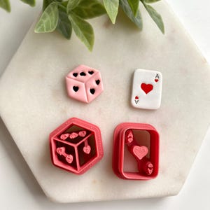 May include: A selection of pink and red heart-themed cookie cutters and a playing card. The dice-shaped cutter has heart-shaped indents. The playing card is white with a red heart and the letter "A".