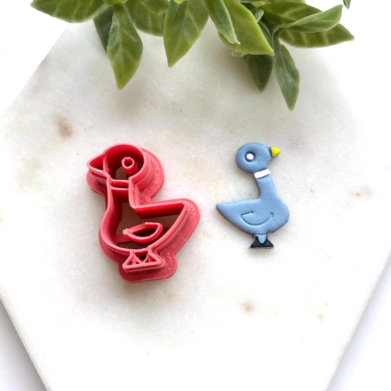 May include: A pink plastic duck-shaped cookie cutter alongside a small, light blue duck-shaped cutout. The cutout features a yellow beak, a white neck band, a white eye, and black feet, mirroring the outline of the pink cookie cutter tool.