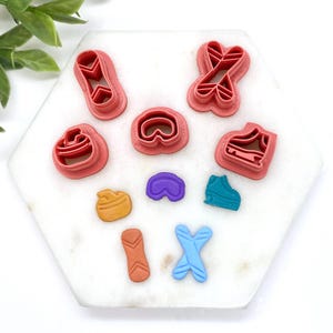 May include: A selection of colourful polymer clay cutters in various shapes, including hearts, shoes, and abstract designs. The cutters are in shades of pink, blue, purple, orange, and yellow, arranged on a white hexagonal surface.