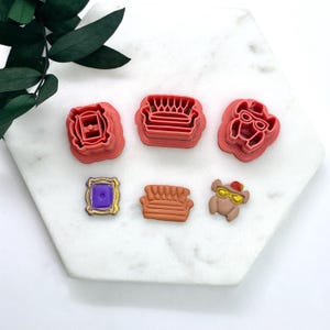 May include: Six red plastic cookie cutters shaped like iconic items from the TV show Friends. The cutters include a couch, a picture frame, a coffee mug, a turkey, and two more couches.