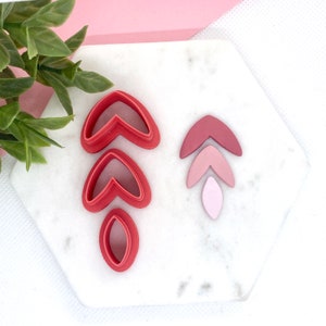 May include: A set of three red heart-shaped cookie cutters and a set of three pink and red leaf-shaped cookie cutters on a white marble surface.
