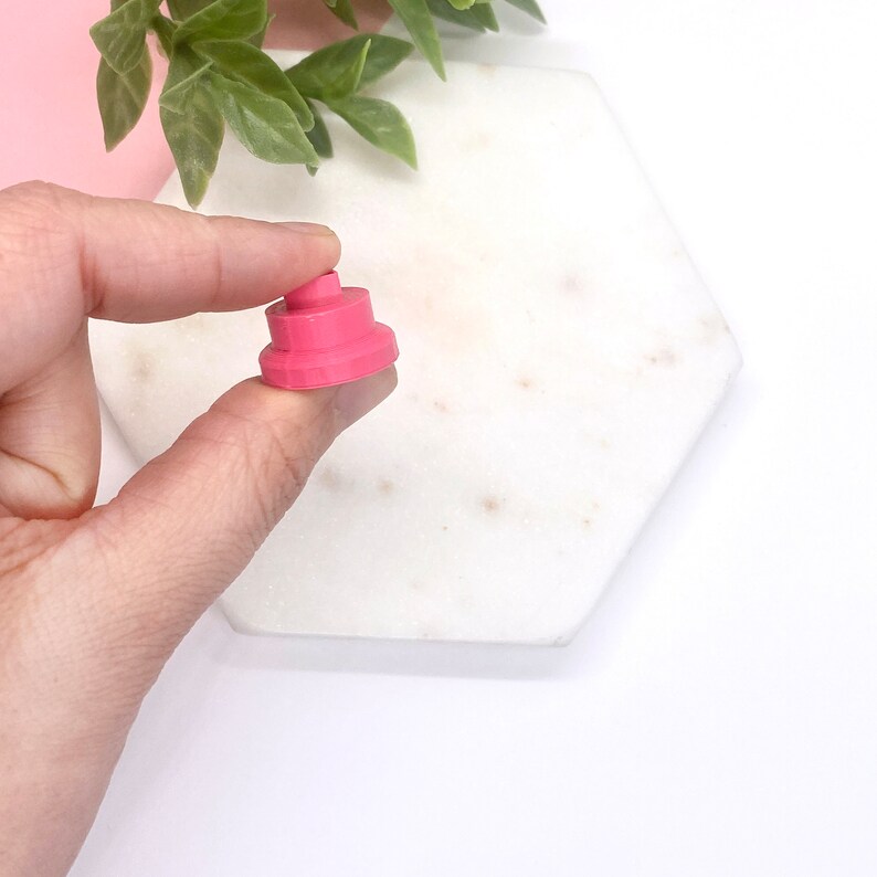 May include: A pink plastic cake topper with three tiers. The topper is small and is being held by a hand.