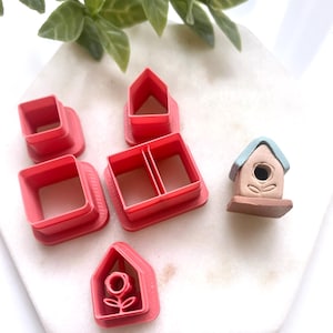 May include: A selection of red plastic cookie cutters in different shapes, including squares, houses, and a house with a flower. A small birdhouse with a blue roof and brown base is also present. The items are arranged on a white surface.