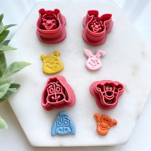 May include: A collection of colorful, edible character shapes, including Winnie the Pooh, Piglet, Eeyore, and Tigger. The shapes are in various colors, such as yellow, pink, blue, and orange. The shapes are on a white surface.