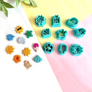 May include: A set of 15 teal blue plastic cookie cutters shaped like various summer and beach themes, including a sun, a seashell, a turtle, a flower, a castle, an ice cream cone, and a wave. The cutters are arranged on a pink and yellow background.